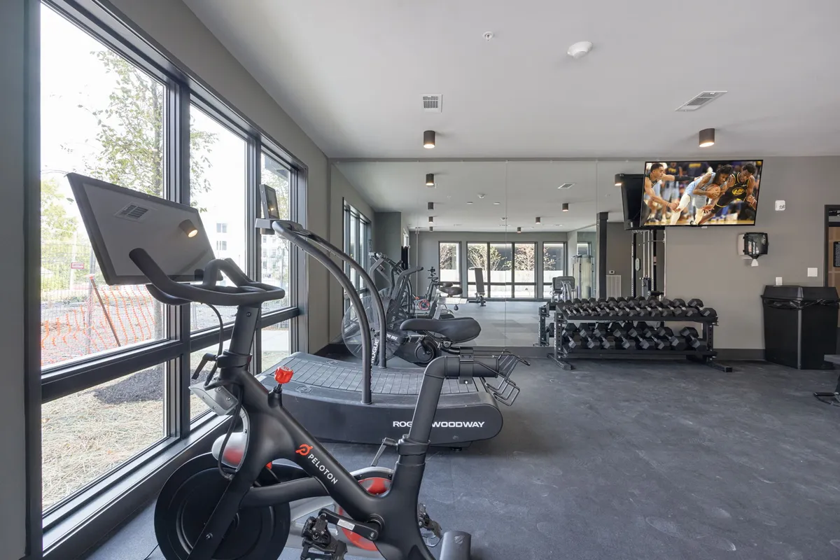 A bright, modern indoor fitness center featuring a Peloton bike, treadmill, free weights, large windows for natural light, and a big TV screen. The space is active, spacious, and invites both individual and group workouts.