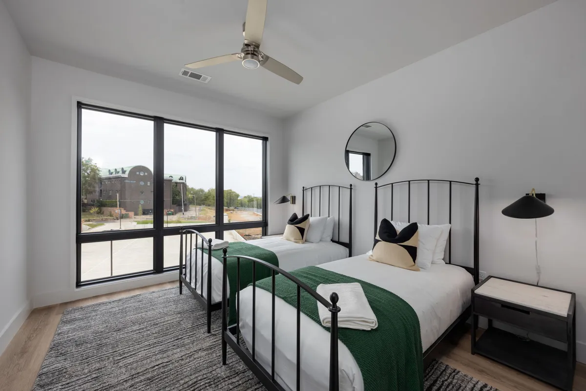 A serene bedroom with two twin beds featuring black metal frames, plush bedding, and modern bedside lamps. Large windows fill the space with soft natural light, and a round mirror above the beds adds a contemporary touch. The room feels comfortable and ideal for guests or family.