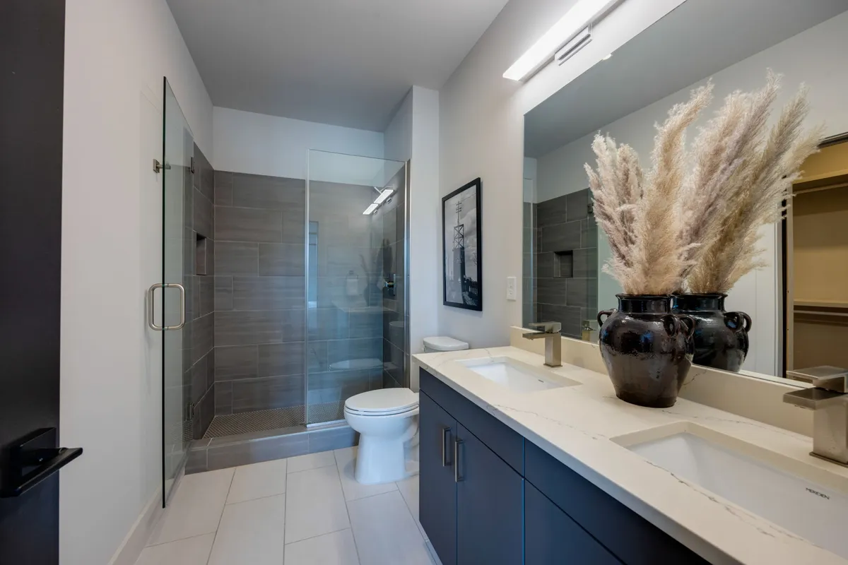 A spa-inspired bathroom with a double vanity featuring modern fixtures, a large glass shower enclosure, decorative pampas grass, and framed artwork. The space exudes calm luxury and is designed for restorative self-care routines.
