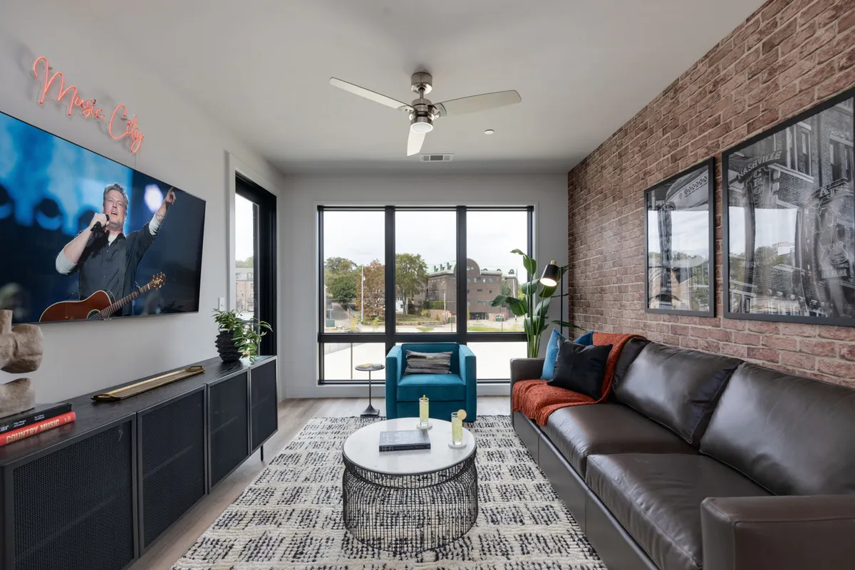 A cozy living room with a brown leather sofa, large windows letting in natural light, a television with a musical performance displayed, and a textured brick wall. The setup is inviting for both relaxation and entertainment. It also visually suggests the presence of a sleeper sofa for extra guests.