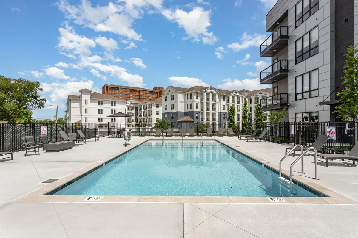 This exterior image captures a vibrant swimming pool surrounded by lounge chairs, landscaped areas, and adjacent buildings. People are seen sunbathing, swimming, and socializing, emphasizing the communal and resort-style vibe.