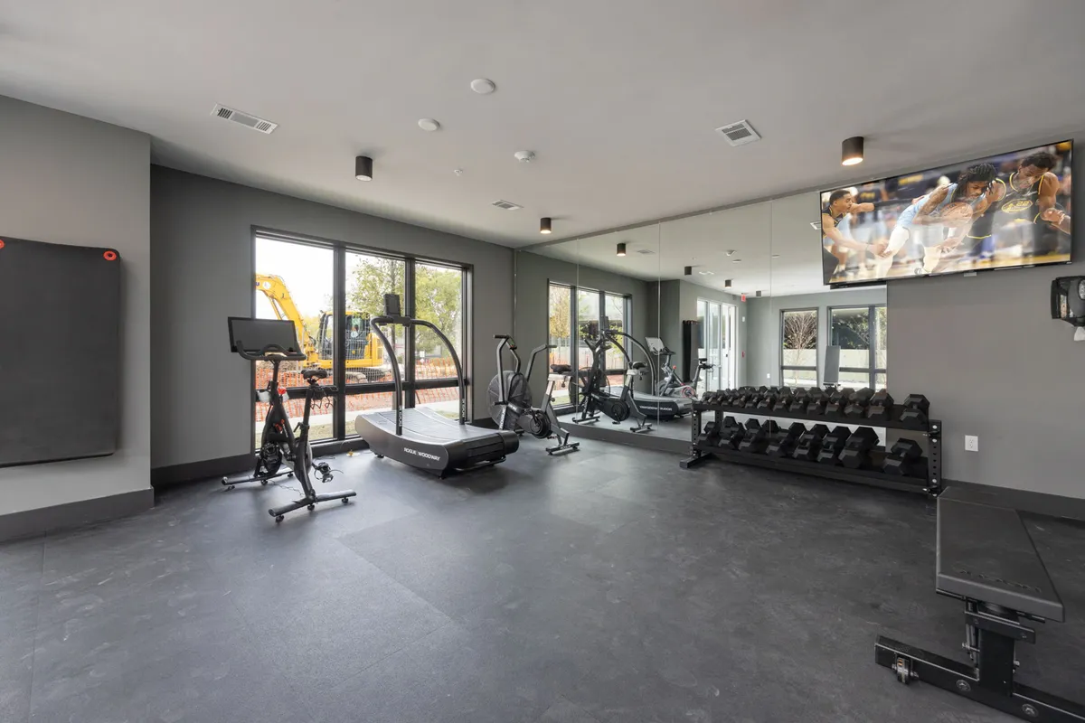A modern fitness center with a treadmill, weight rack, exercise bike, large mirror, and wall-mounted television. The space feels bright, open, and energetic—ideal for maintaining an active lifestyle.