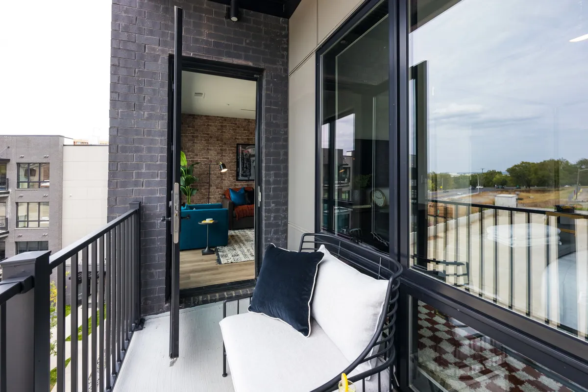 A stylish balcony features comfortable seating, contemporary planters, and a view into the interior living space, suggesting a smooth transition between indoor and outdoor living. The mood is urban and relaxed—ideal for morning coffee or sunset toasts.