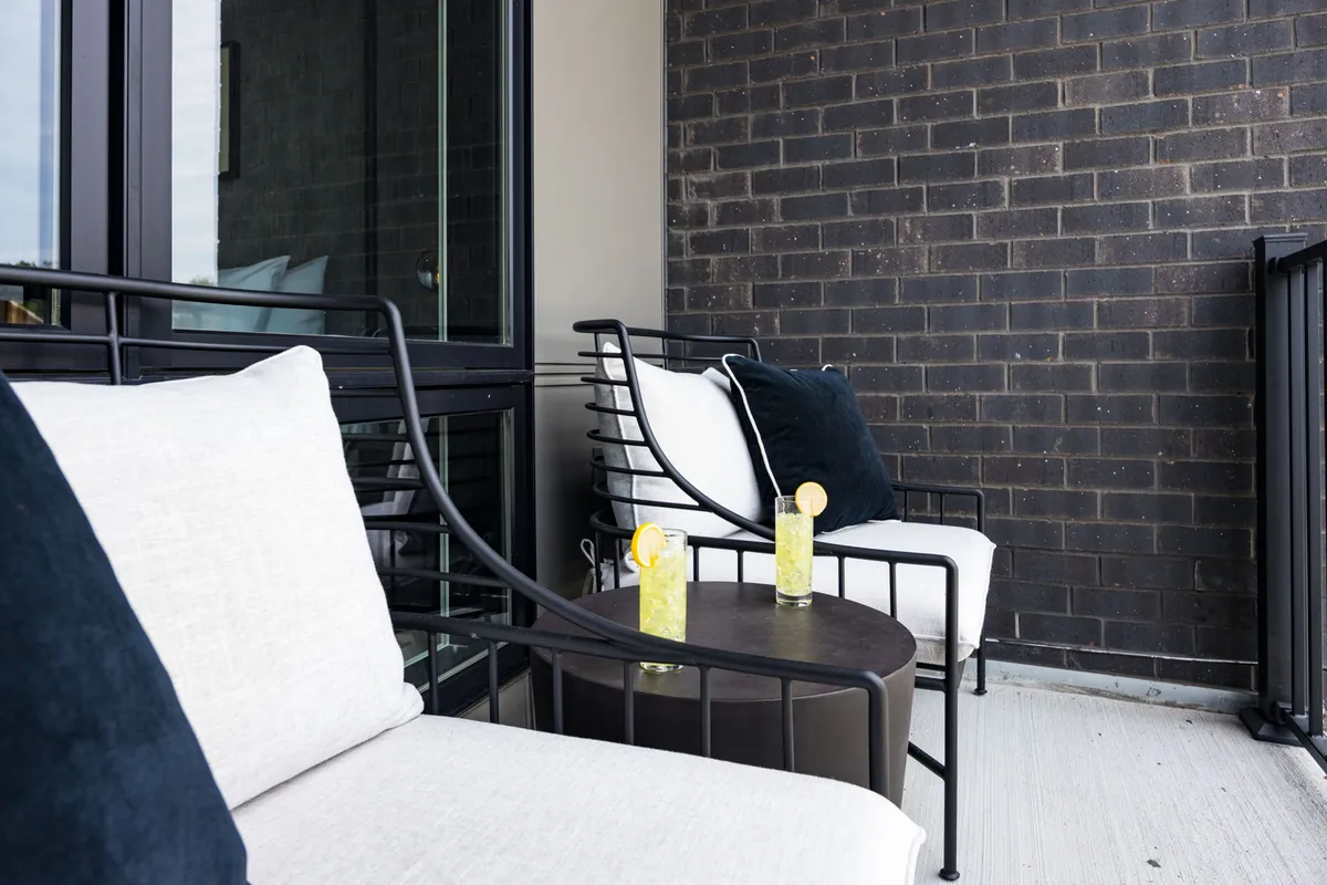 A cozy balcony setup with stylish chairs and a small round table, perfect for enjoying drinks or morning coffee. The backdrop features a classic brick wall and city views, offering a sense of privacy and comfort while still being connected to the urban environment.