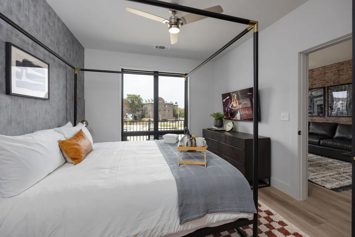 A spacious, stylish bedroom with a four-poster king bed, large windows streaming in natural light, a smart TV atop a sleek dresser, and a modern nightstand. The room is designed with comfort and relaxation in mind, highlighted by plush bedding and a tranquil color palette.
