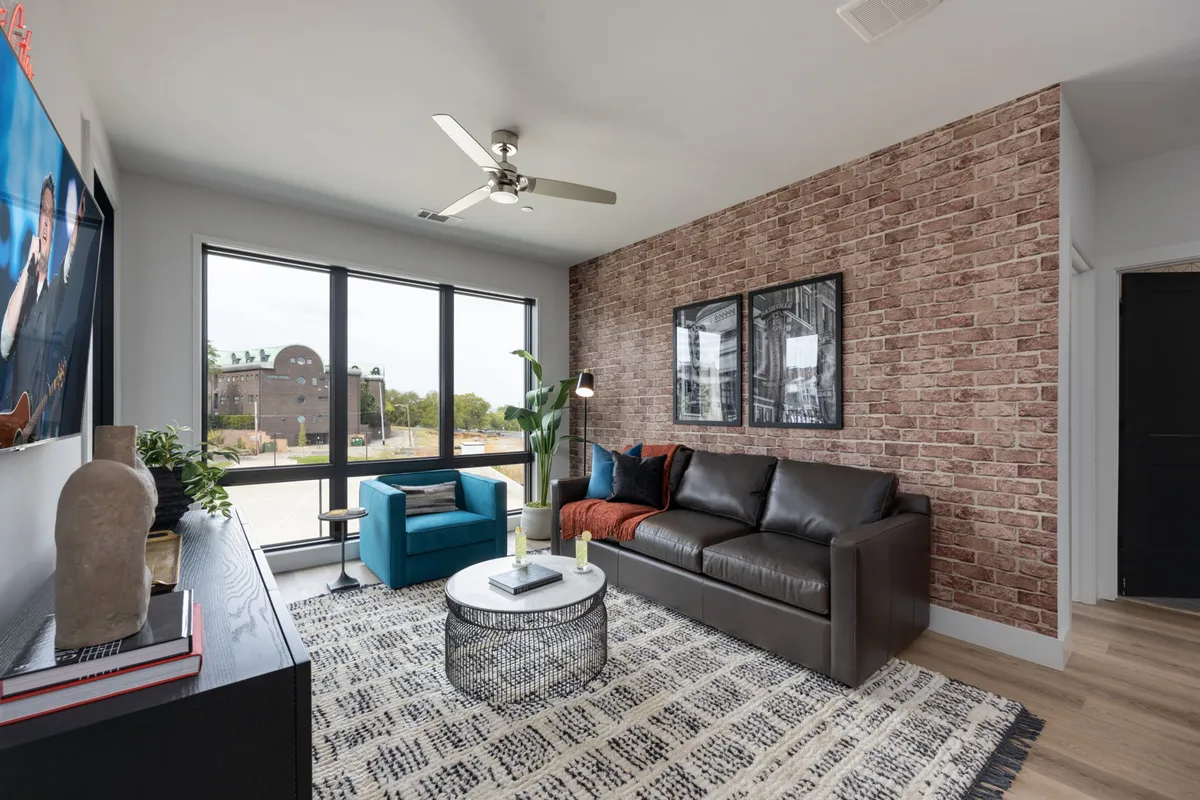The image showcases a stylish, sunlit living room with a plush modern sofa and a vibrant accent chair, framed by expansive windows that flood the space with natural light. Eye-catching artwork adorns the wall, and the scene evokes a sense of comfort and sophisticated relaxation—perfect for unwinding or gathering with friends.