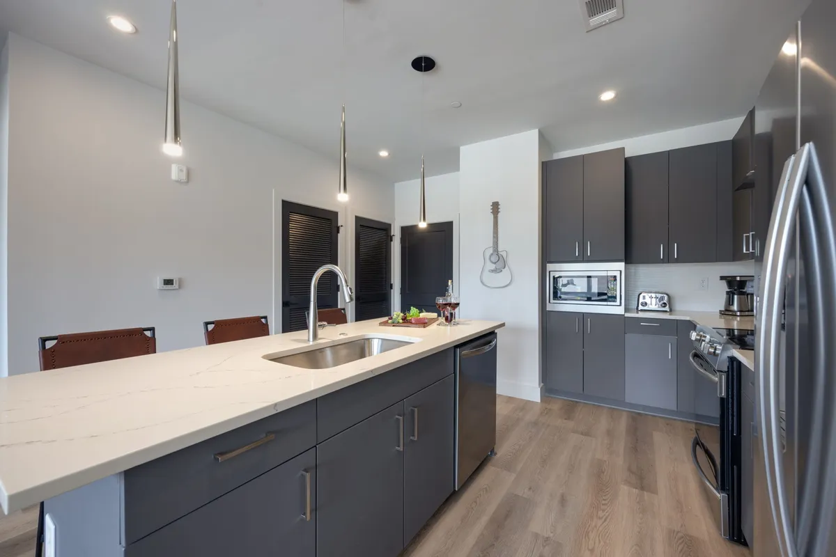 A bright, contemporary kitchen scene centered on a large island with a sink, surrounded by stainless steel appliances, a guitar on the wall, and fresh produce on the counter. The open-plan design suggests space for family gatherings and social cooking sessions.